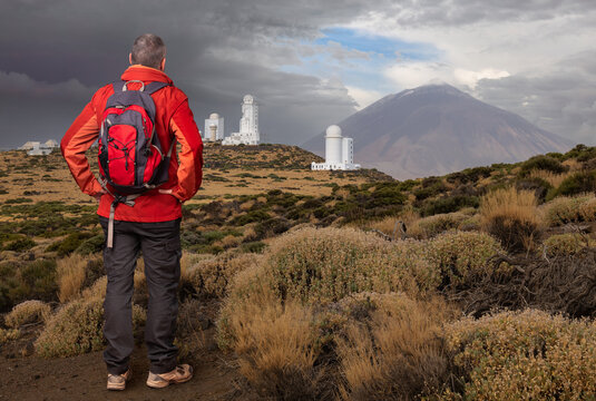 A hiker stands in the landscape of the volcanic island of Tenerife and looks at the white buildings of the observatory and the Teide volcanic cone. There are bushes and grasses everywhere.