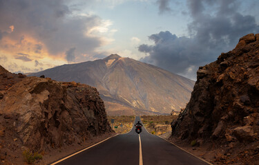 A motorcyclist has passed through a gorge and is driving on the straight and hilly road towards Mount Teide on the island of Tenerife. It is just before sunset in the evening.