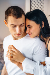 Attractive, hot, sexy brunette sits high on the kitchen furniture. Hugging boyfriend with legs passionately. Woman presses her body to the man. Dressed in white shirts and dark jeans