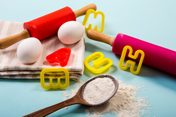 Cooking class for kids concept. Eggs, flour, rolling pins, letter shape cookie cutters on light blue background, studio shot.