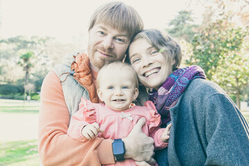 Happy family couple and little kid posing outdoors. Parents holding cute baby girl in arms, standing in autumn park, looking at camera. Medium shot. Family portrait concept
