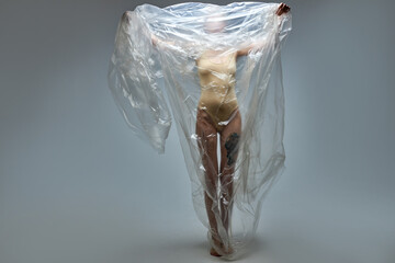 weak tired slim model woman with plastic bag on body, posing at camera isolated on gray studio background. Plastic pollution problem and environment protection concept