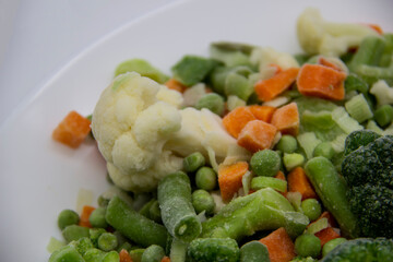 Frozen vegetables: cauliflower, green peas, leeks, broccoli, carrots, green beans on a white plate.