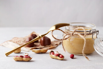 Glass jar with natural peanut butter, golden spoon, peanuts and sweets on a light background. Delicious breakfast close-up. Copy space