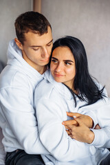 Beautiful young couple in the same white hoodie and black pants lie on the bed hugging. Dreaming of happy future together. Enjoying each other's company. Man gently hugs his beloved from back