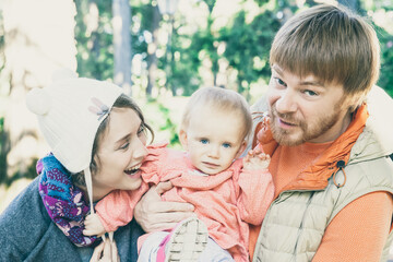 Fototapeta premium Joyful parents and toddler daughter having fun while walking outdoors, posing and laughing. Dad holding kid, mom wearing funny girls hat. Family portrait concept