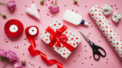 Obraz na płótnie Canvas Valentine's day gift wrapping concept.Box wrapped in white paper in hearts,red bow,confetti,red ribbon,roses,cotton, glue,scissors and a roll of wrapping paper on a pink background,top view,flat lay.