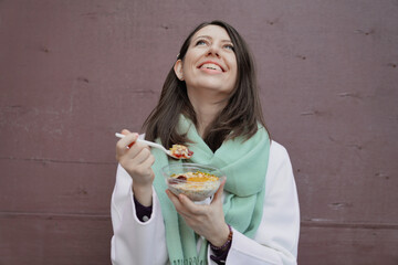 Attractive brown hair young girl in white coat and scarf having breakfast, lunch to-go, cheerful, smiling. Plain wall on background, copy space for text