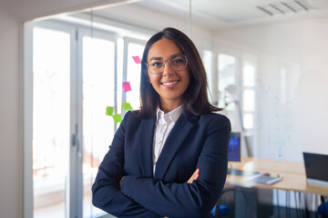 Confident Latin business leader portrait. Young businesswoman in suit and glasses posing with arms folded, looking at camera and smiling. Female leadership concept - obrazy, fototapety, plakaty