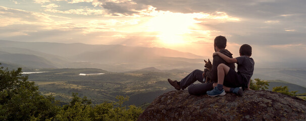 Father, son and dog on top of rock against sunset over Rhodope mountains, Bulgaria. Family spends...