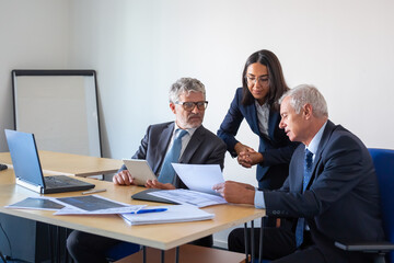 Serious colleagues reading document and working with statistics. Professional content businesspeople in office suits sitting at table with laptop, papers and tablet. Management and partnership concept