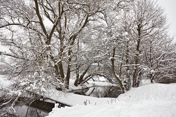 Small river in winter. Rural January scene, bridge over creek. Trees on riverbank covered with snow.