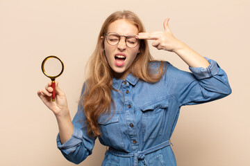blonde woman looking unhappy and stressed, suicide gesture making gun sign with hand, pointing to...