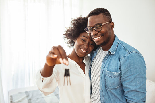 Happy Young Family Couple Holding Key To New Home On Moving Day Concept, First Time Real Estate Owners Man Husband Embrace Woman Wife Look At Camera Proud Buying Property Stand In Own Flat With Boxes