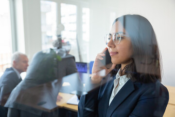 Confident office professional in suit and glasses talking on cellphone in office of her boss behind glass wall. Copy space. Communication and advertising concept