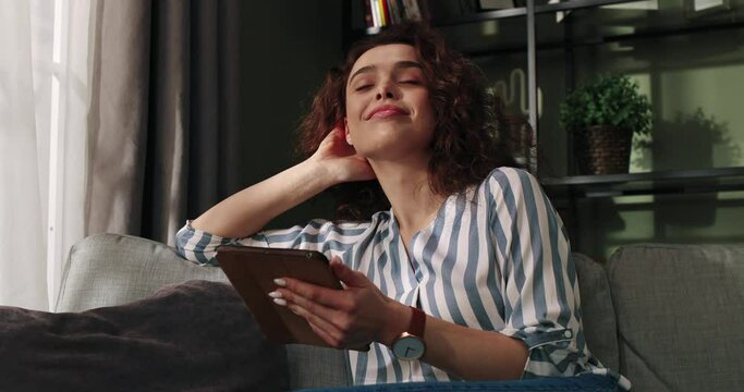 Close up of happy Caucasian cheerful young female smiling in good mood sitting in apartment alone typing on tablet device surfing internet with dreamy face, positive emotions, Leisure concept
