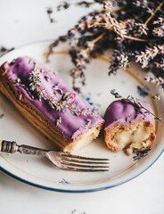 Lavender eclair with cream decorated with purple flowers and vintage fork on oval plate over white marble background, selective focus, close-up. Modern French style sweet gourmet dessert concept
