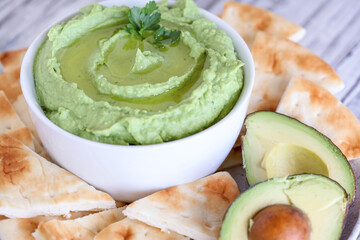 Vegan avocado Hummus, made with chickpeas, avocados and tahini, with olive oil. Garnished with parsley and served with pita bread over a rustic table. Extreme shallow depth of field with background.