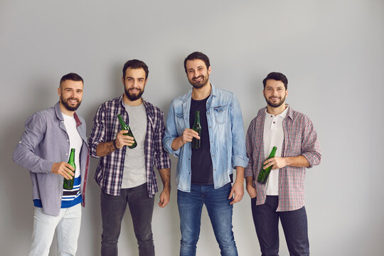 Young people enjoying alcoholic drinks. Happy friends drinking beer and having good time. Portrait of 4 men standing in studio with light background, holding green glass bottles and smiling at camera