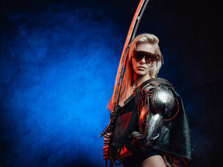 Gorgeous and seductive female warrior with blond hairs and sword from the future poses in dark background.