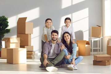 Fototapeta na wymiar Portrait of happy smiling young family with little children sitting on floor in new home with unpacked boxes. Concept of moving day, real estate, mortgage, buying house or apartment