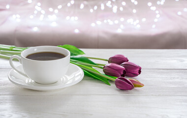 Obraz na płótnie Canvas A cup of coffee and tulips on a light background with burning lights. Side view, with space to copy. The concept of holiday backgrounds.
