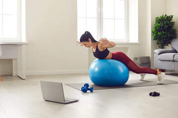 Sporty healthy lifestyle. Slender and sporty woman in sportswear trains at home in the morning with a fitness ball and watches her favorite show on a laptop. Home workout concept.