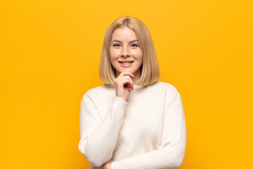blonde woman looking happy and smiling with hand on chin, wondering or asking a question, comparing...