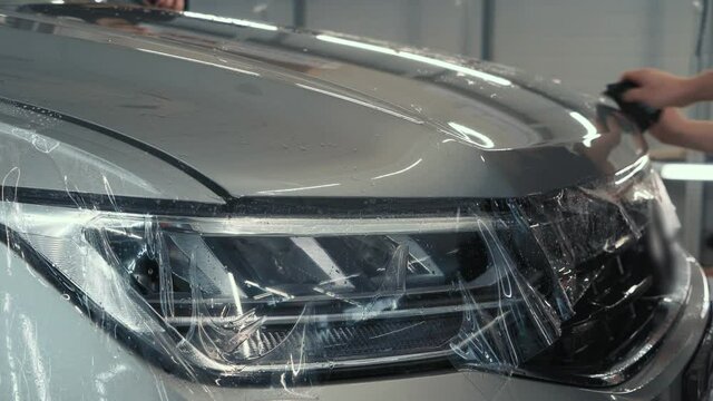 Workers wrap wet Paint Protection Film or anti-gravel protection coating on new car hood. Car detailing.