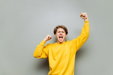Portrait of a joyful young man in yellow emotional clothes rejoices in victory on a gray...