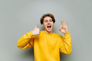 Joyful guy stands against the gray wall and looks at the camera with a happy face and smiles, shows...