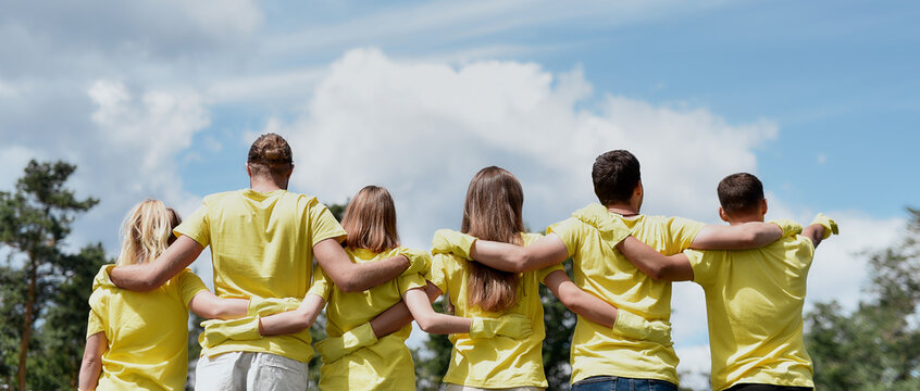 Love your nature. Group of young volunteers wearing uniform and rubber gloves hugging and looking at green forest in front of them, rear view