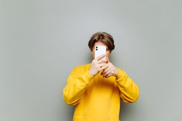 Young man in a yellow sweatshirt covers his face with a smartphone on a gray background. Guy uses...