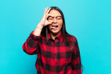 latin woman smiling happily with funny face, joking and looking through peephole, spying on secrets