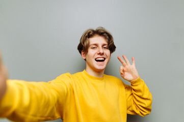 Positive guy takes a selfie against a gray wall and shows OK gesture with his fingers, looks at the...