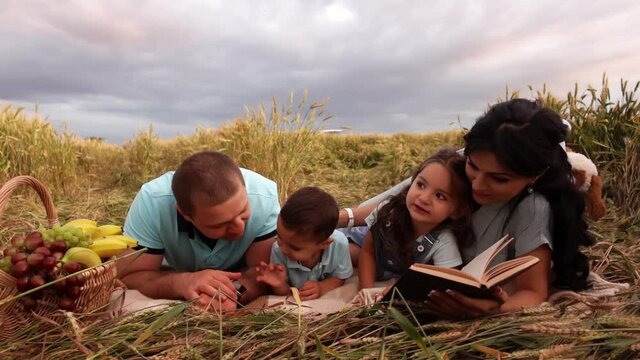 Family is laying at the blanket on picnic with books and mom reads aloud. Father kisses his son. The conception of happy together vacation and travelling outdoors.