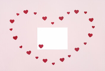 Greeting card for Valentine's Day. Romantic composition with red paper hearts and a blank card on a pink background. Copy space, top view