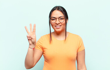 latin woman smiling and looking friendly, showing number three or third with hand forward, counting...