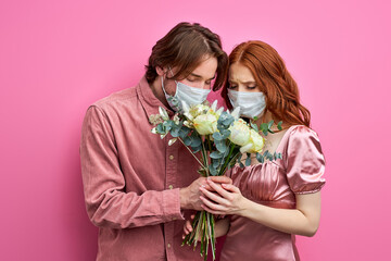 man and woman in medical masks trying to smell the scent of flowers, holding bouquet in hands. loss of smell due to coronavirus, covid-19. isolated pink background