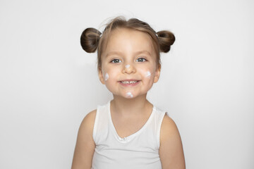 Kid cosmetics, face cream, skin care concept. Adorable smiling little girl with cream on her face, copying mom behavior, using cosmetics. Isolated on white, copy space