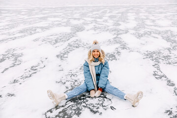 Cheerful young blonde woman, sitting on ice on frozen lake, on a cold winter day, smiling, looking at camera.