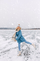 Cheerful young blonde woman skating on ice on a frozen lake on a cold winter day in snow fall.