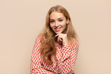 blonde woman looking happy and smiling with hand on chin, wondering or asking a question, comparing...