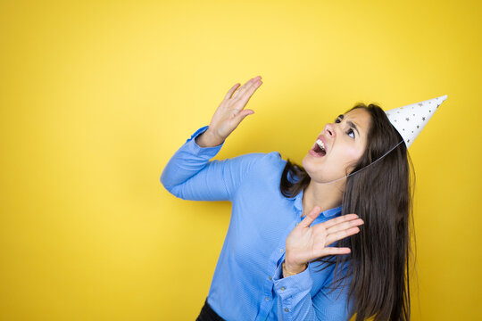 Young Caucasian Woman Wearing A Birthday Hat Over Isolated Yellow Background Scared With Her Arms Up Like Something Falling From Above