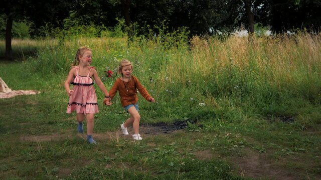 Two hippie girls hold hand and run together on green grass. Children in boho style are resting in nature.