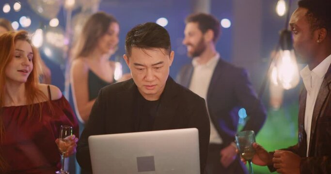 Chinese workaholic young adult man performing online project on computer laptop while walking between colleagues dancing celebrating corporate party.
