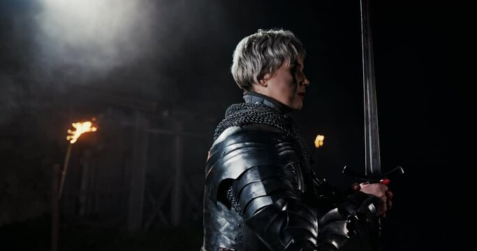 A brave female warrior with a sword in her hands stands on the battlefield in the dark with a peek of torches and is ready to strike the brutal sword looking the enemy in the eye.