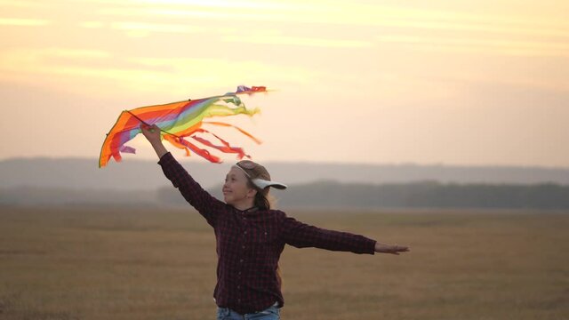 Girl wants to become a pilot. Child dreams of freedom, flight and travel. Happy child in pilot's helmet runs with multi-colored kite in his hands at sunset. Teenager playing on a plane outdoors.