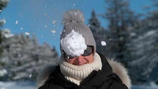 SLOW MOTION, PORTRAIT, CLOSE UP, DOF: Smiling female tourist on carefree winter vacation gets hit in the face by a fluffy snowball. Cheerful young Caucasian woman gets involved in a fun snowfight.