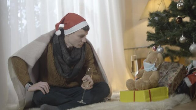 Depressed young man drinking champagne with toy teddy bear in Covid face mask at home. Portrait of frustrated isolated Caucasian guy celebrating lonely Christmas indoors. Coronavirus pandemic holidays